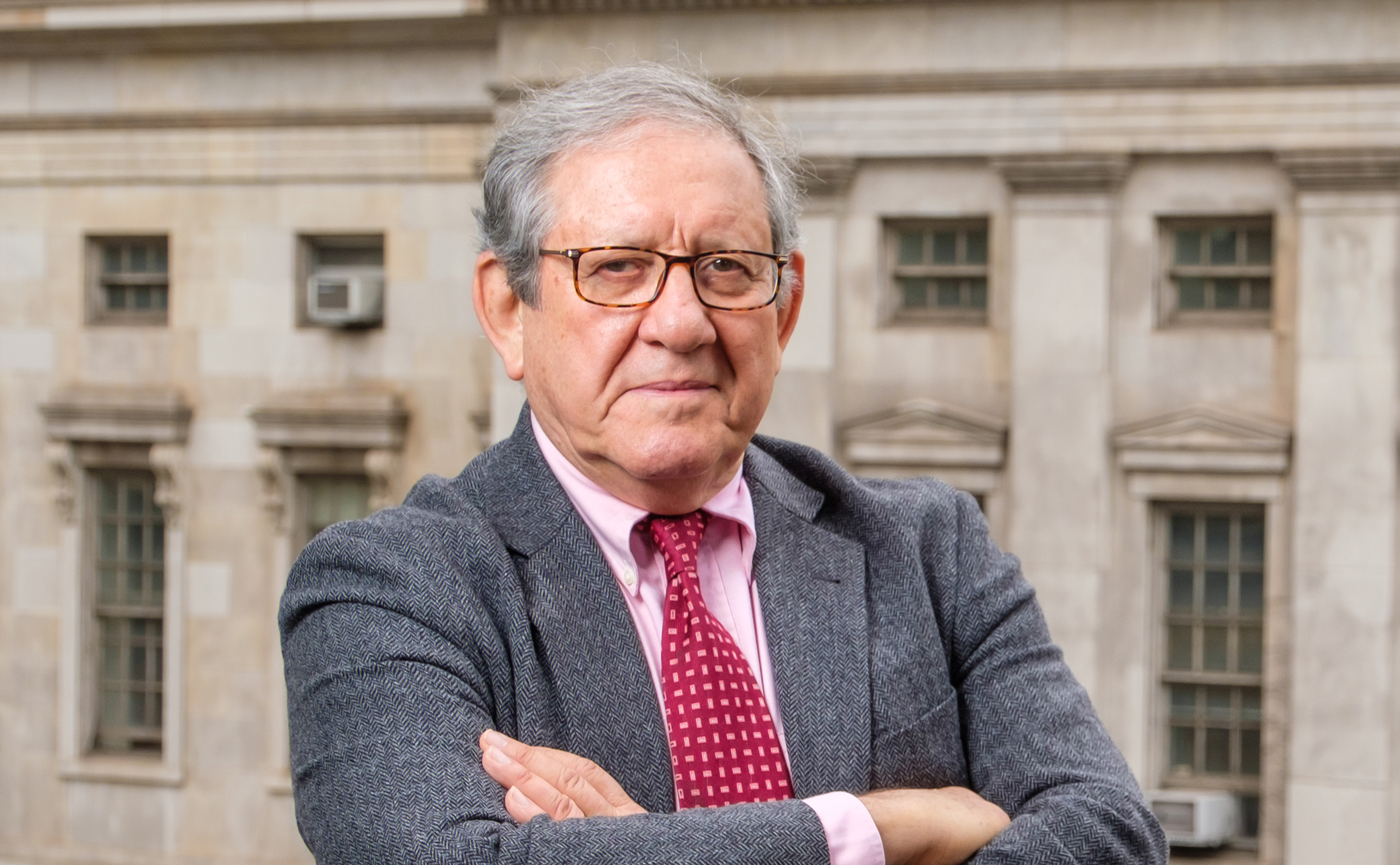 A man in a gray suit and red tie posing in front of a stone building with his arms crossed.