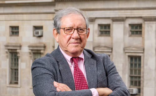 A man in a gray suit and red tie posing in front of a stone building with his arms crossed.