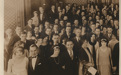 An old photo from the 1920s featuring dozens of fancily dressed people in an ornate room.
