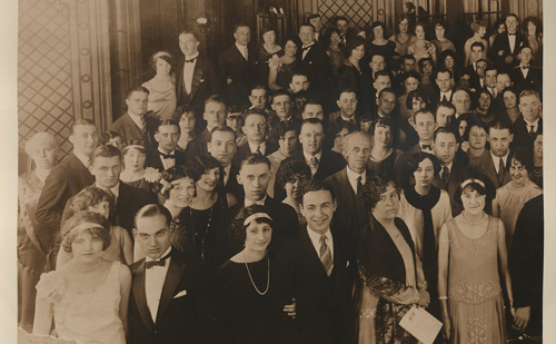 An old photo from the 1920s featuring dozens of fancily dressed people in an ornate room.