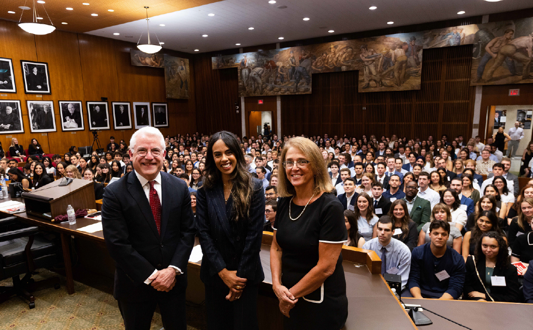 A man and two women, formally dressed, in a courtroom with a full room of incoming students behind them.