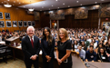 A man and two women, formally dressed, in a courtroom with a full room of incoming students behind them.
