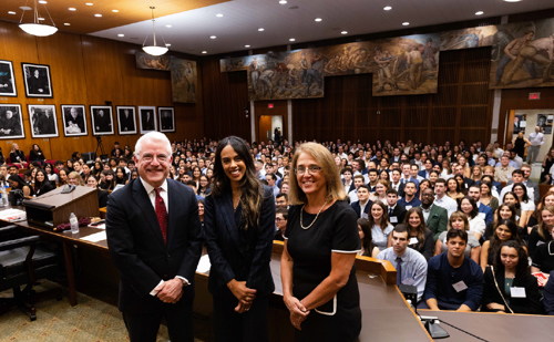 A man and two women, formally dressed, in a courtroom with a full room of incoming students behind them.