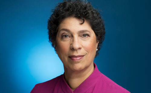 A woman with short, wavy black hair wearing a pink blouse poses in front of a blue background.
