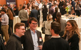 Three young men and one young woman mingle in the foreground of a banquet setting with many people and fancy tables in the background.