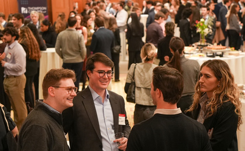 Three young men and one young woman mingle in the foreground of a banquet setting with many people and fancy tables in the background.