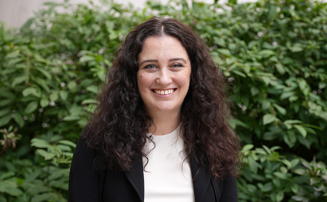 A woman with curly dark hair smiles in front of a bush wearing a white shirt and black jacket.
