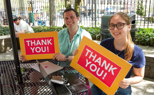 A man and a woman sitting at a black table outdoors holding signs that say "Thank You!"
