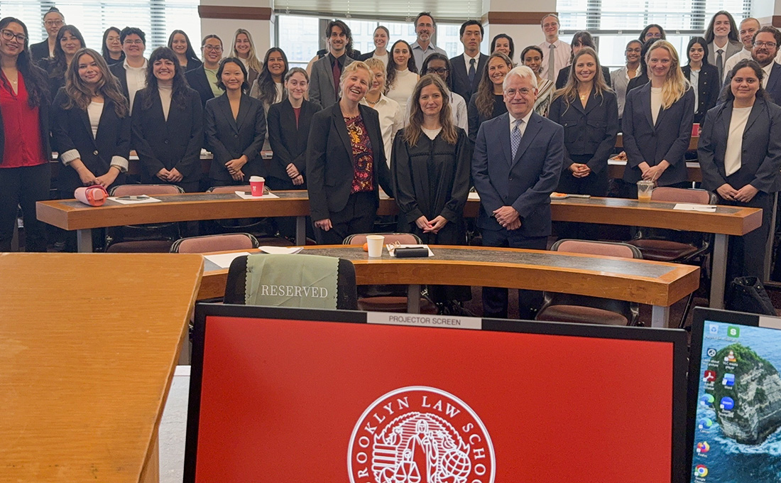 A couple dozen people pose in a classroom with a monitor depicting the Brooklyn Law School shield front and center.