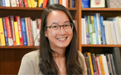 A woman with long hair and glasses smiling in front of a bookcase.