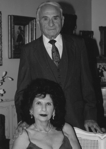 A black-and-white photo of an older man in suit and tie standing behind a woman sitting in a chair.