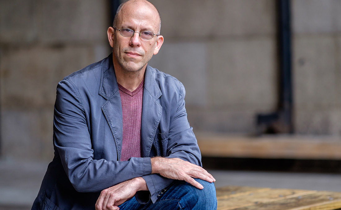 A man wearing glasses in jeans and a blazer poses while leaning with his hands on his knee.