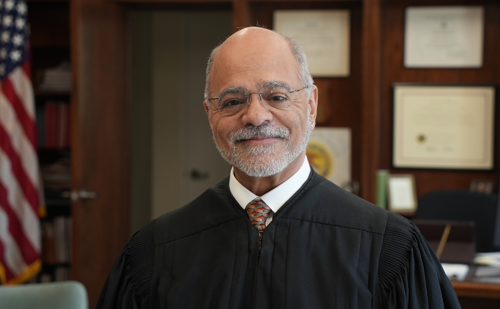 A bearded man in judge's robes poses in an office.