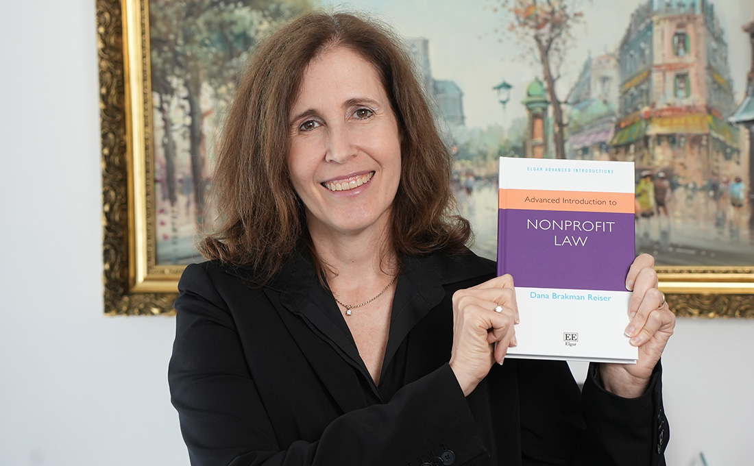A woman in a blazer poses in front of a painting holding up a book titled "Advanced Introduction to Nonprofit Law."