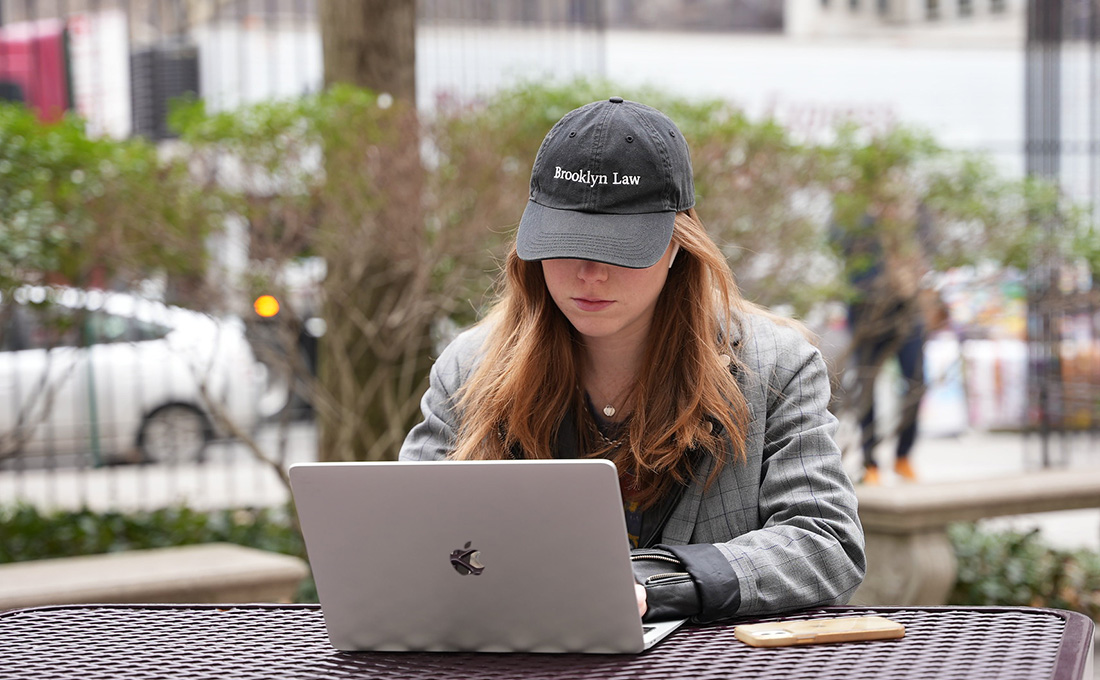 A female student with a Brooklyn Law hat working on a laptop outside.