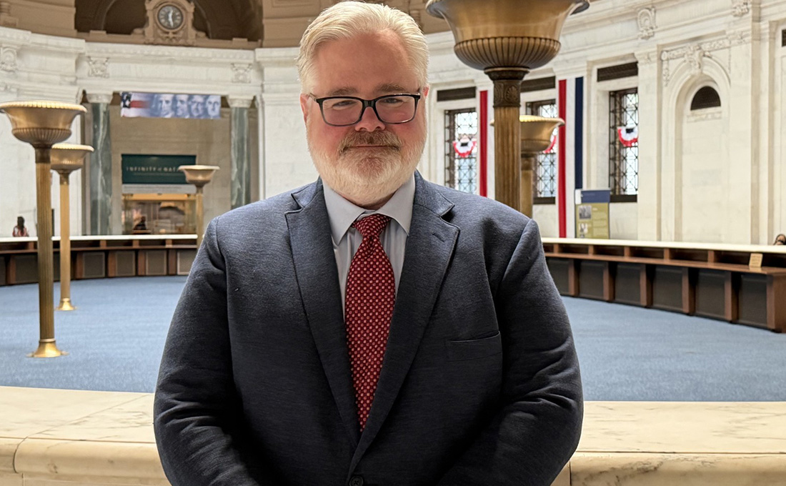 A man in a suit with a beard and glasses poses inside of a large marble room.