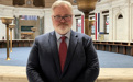 A man in a suit with a beard and glasses poses inside of a large marble room.