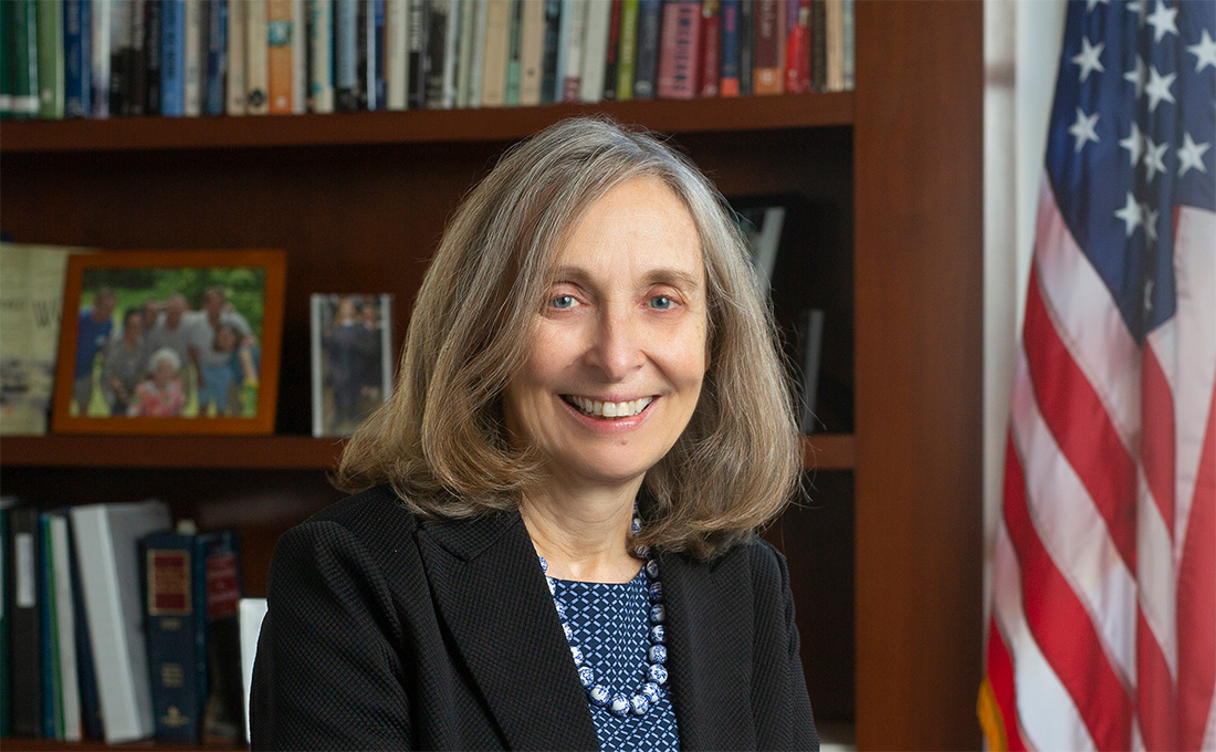 A woman posing in her office with a wooden bookshelf behind her full of books and photos and an American flag next to it.