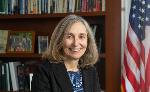 A woman posing in her office with a wooden bookshelf behind her full of books and photos and an American flag next to it.