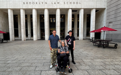Three people pose in front of Brooklyn Law School's main entrance, with two men standing to the side of a woman in a wheelchair.