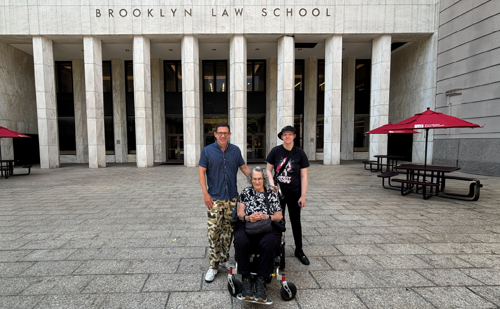 Three people pose in front of Brooklyn Law School's main entrance, with two men standing to the side of a woman in a wheelchair.