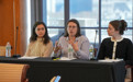 Three women seated at a table with microphones and notepads in front of them.