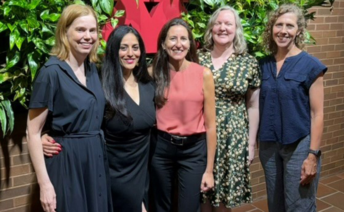 Five women pose in front of a brick wall with decorative plants.