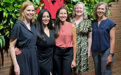 Five women pose in front of a brick wall with decorative plants.