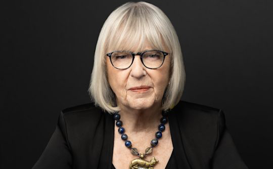 A woman in a black suit poses in front of a black background.