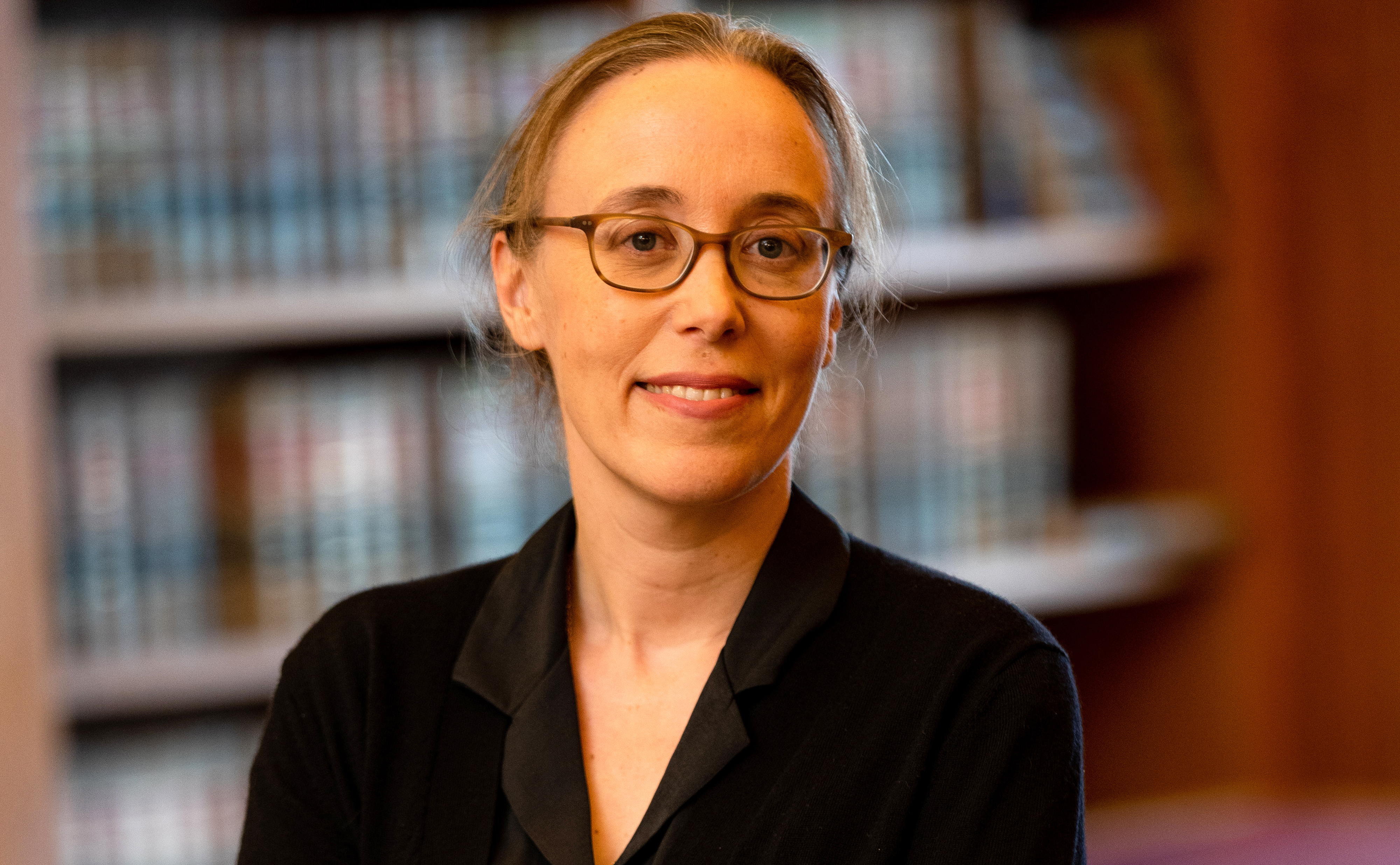 A woman with glasses in front of a bookcase.
